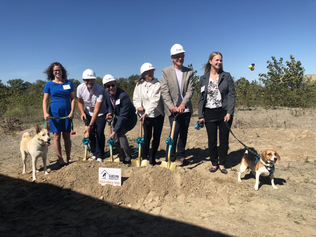 Group in hard hats holding shovels with two dogs at a groundbreaking on a dirt lot