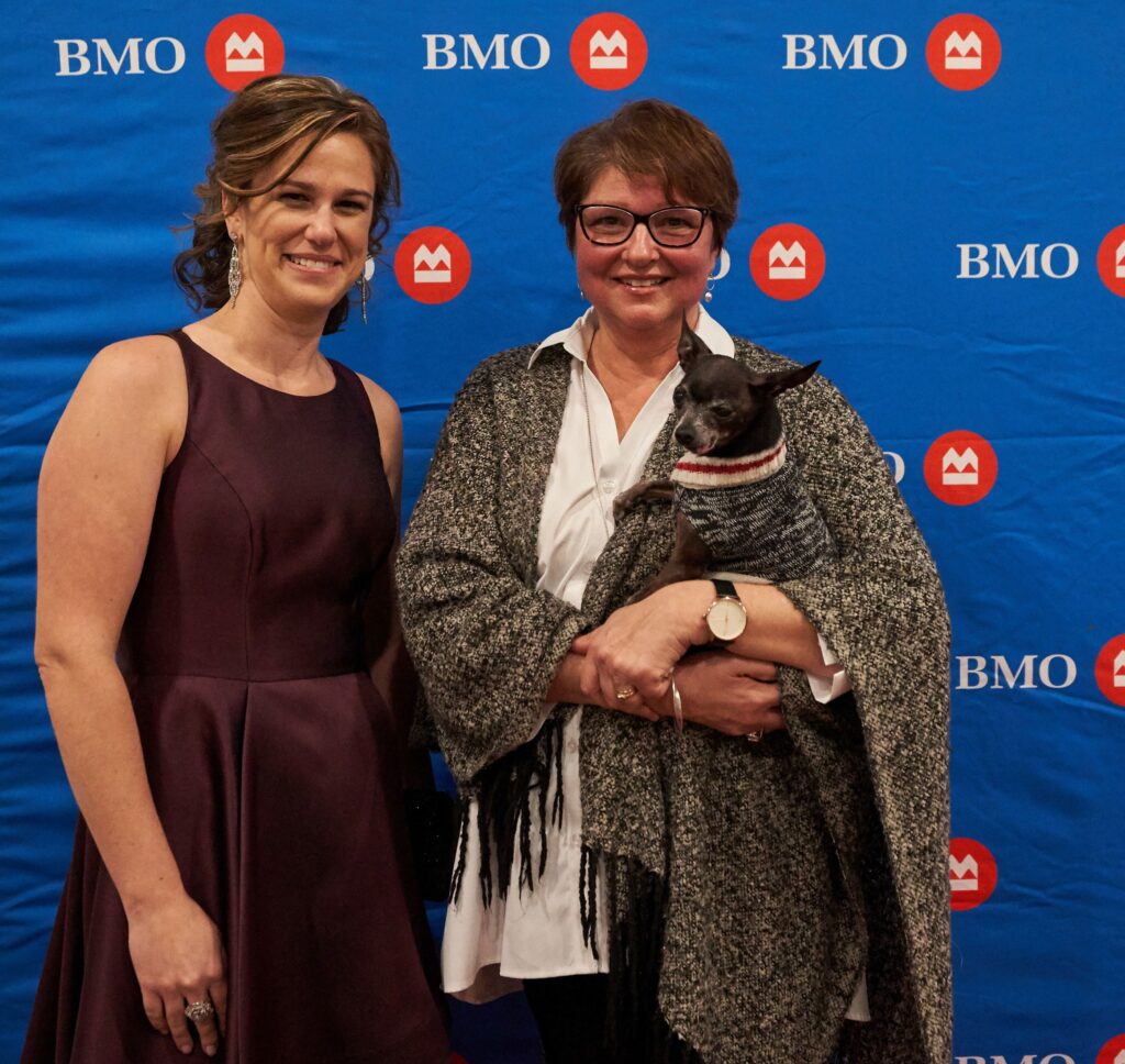 Two women posing in front of a BMO backdrop at the gala, one holding a small black dog