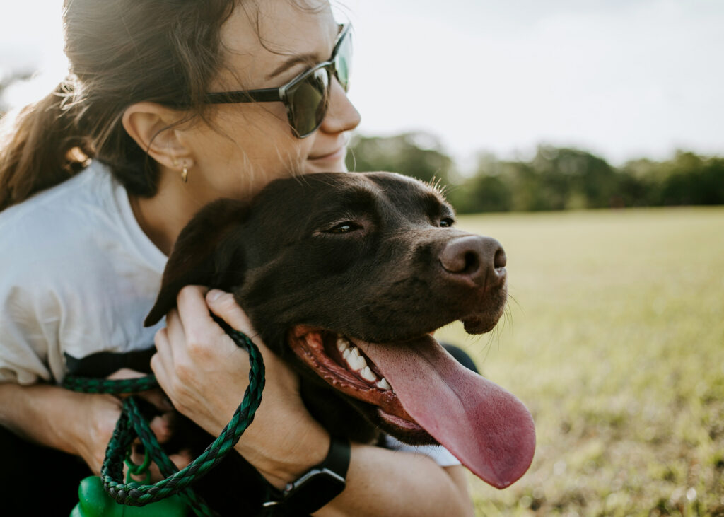 A woman in sunglasses crouches in a field with her arms around a black labrador, which pants happily.