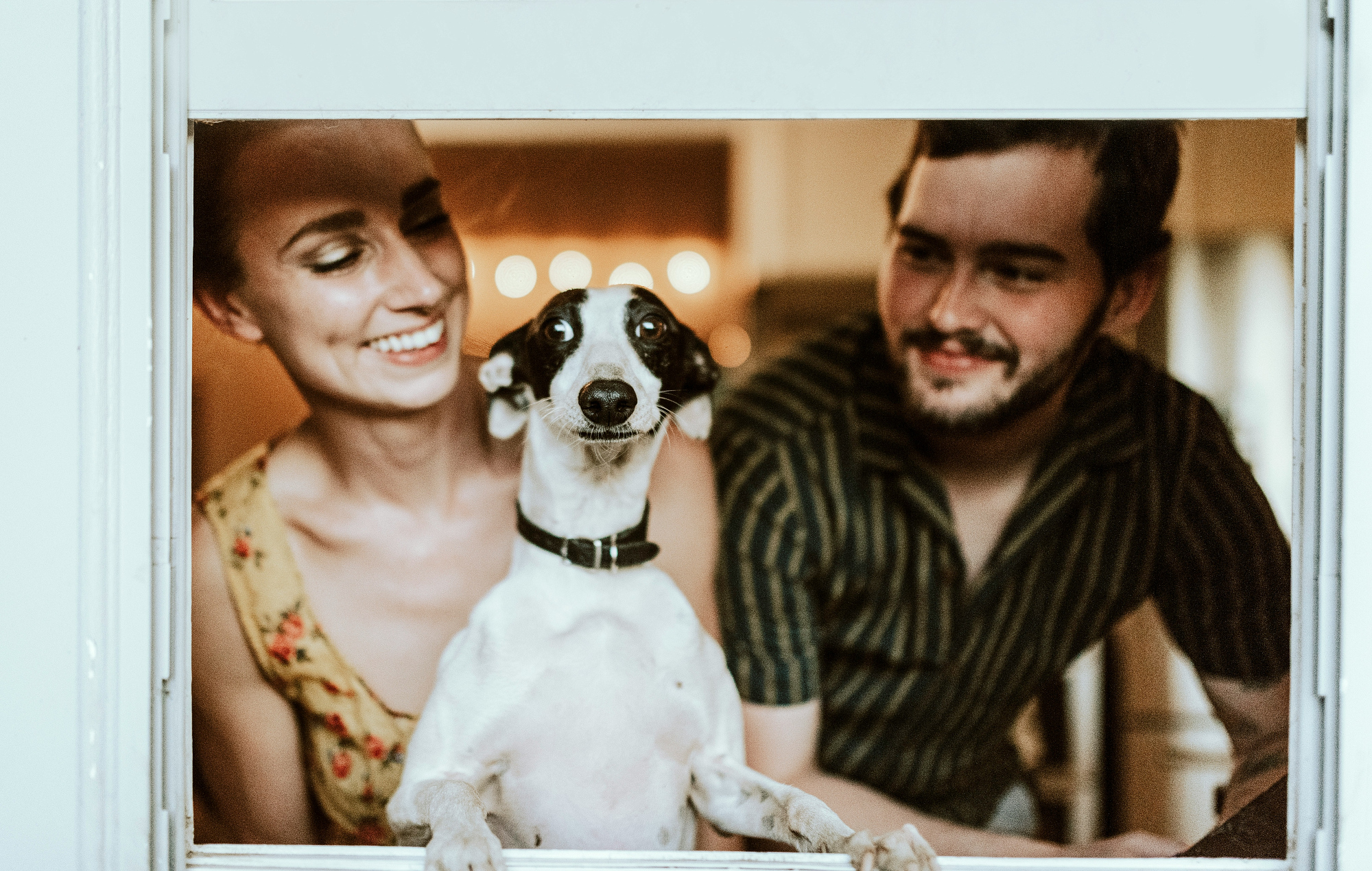 A couple smiles down at a small black and white dog held up between them, framed through a window.
