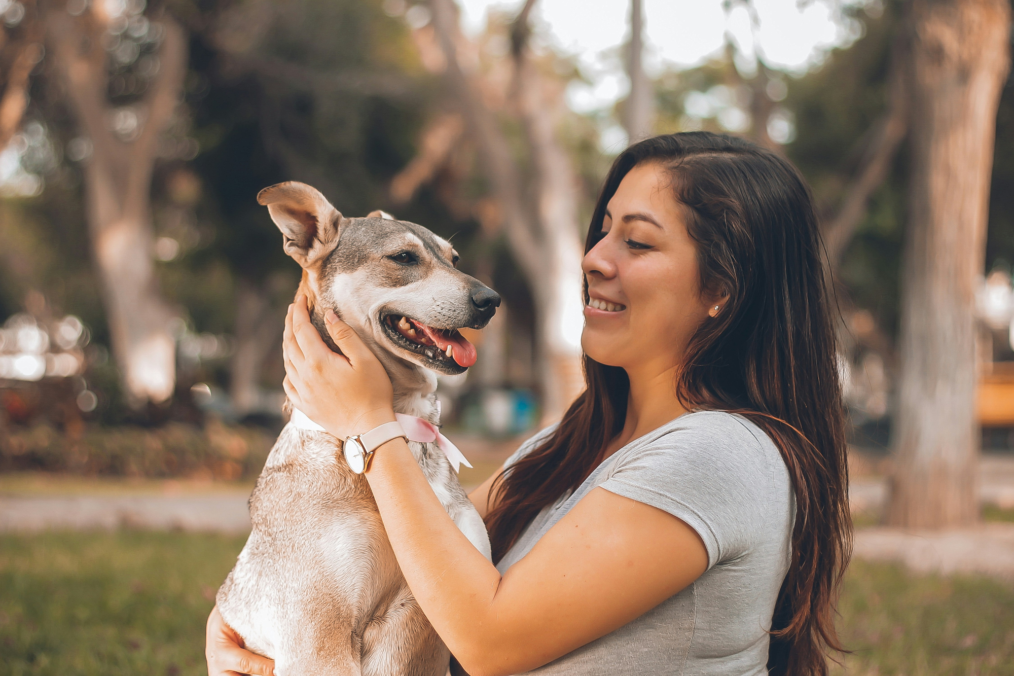 A woman holds a tan dog up in her arms in a park, both looking happy.