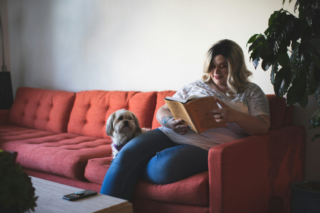 A woman reads a book on an orange sofa with a small shih tzu resting against her leg.