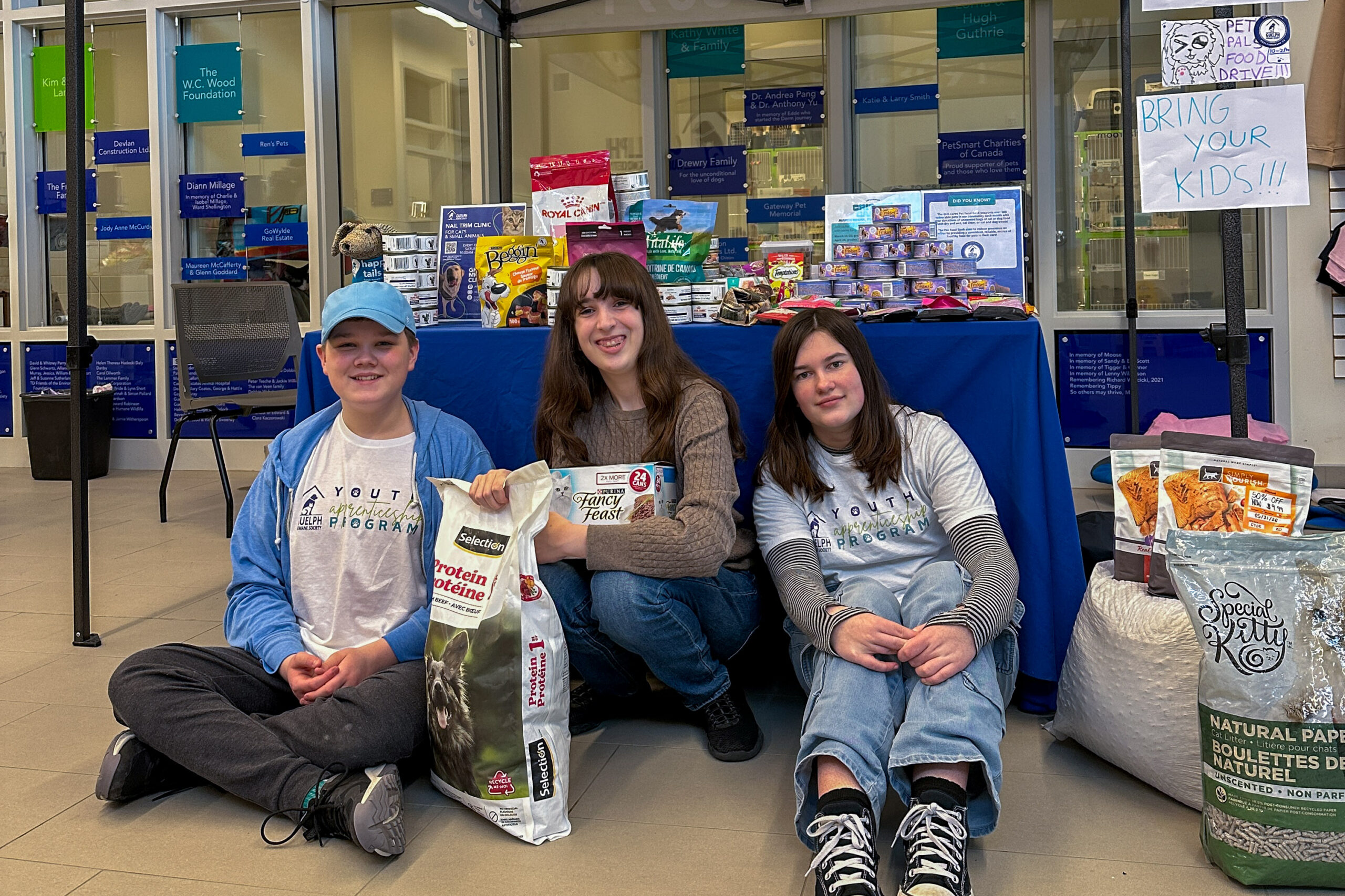 Three youth volunteers sitting on the floor with bags of donated pet food in front of a display of pet supplies